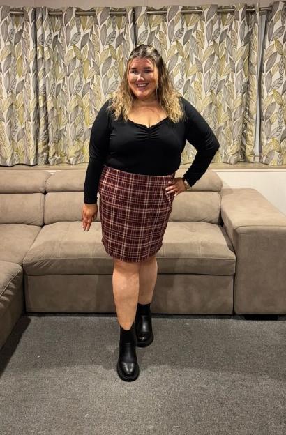 Becki Jones smiling, wearing a black top, plaid skirt, and boots, standing in a room with a beige sofa and leaf-patterned curtains.