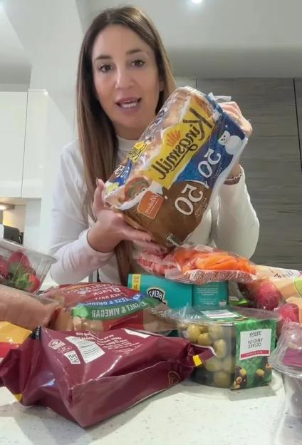 A woman holds up a package of bread in a kitchen with groceries spread across the counter.