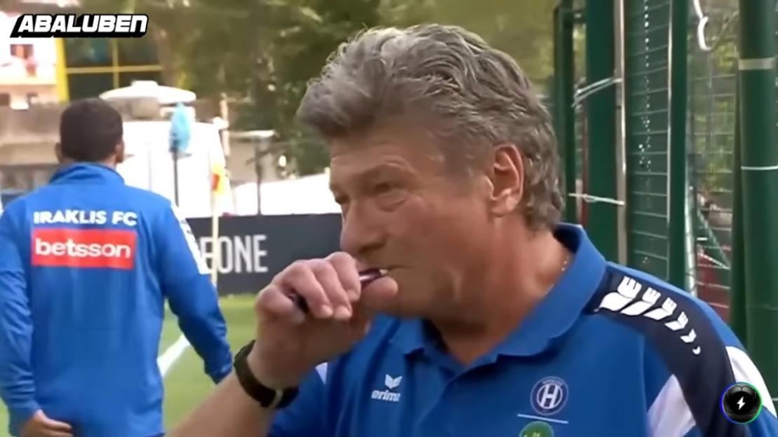 A man in a blue polo shirt chewing on a purple object, with another man in a blue jacket with "IRAKLIS FC betsson" on his back in the background.