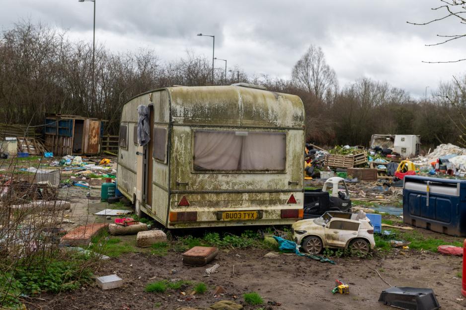 An abandoned, moldy caravan surrounded by litter, broken toys, and waste on a muddy plot of land.