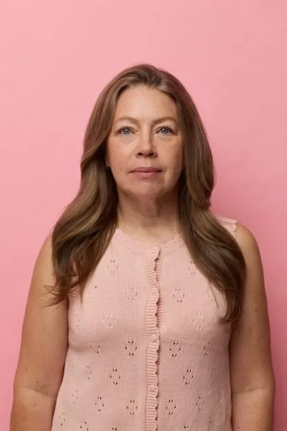 A woman with long brown hair, wearing a pink sleeveless button-down top, stands against a solid pink background.
