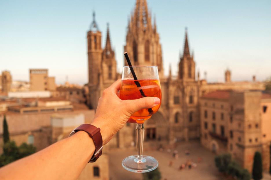 A hand holds a glass of orange Spritz against the backdrop of Barcelona Cathedral’s Gothic spires.