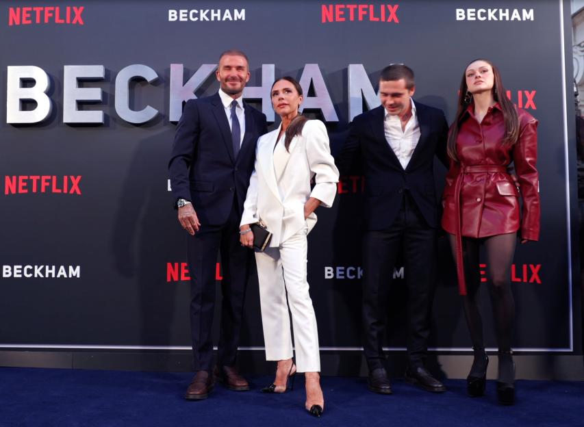 David Beckham, Victoria Beckham, Brooklyn Beckham, and Nicola Peltz Beckham pose together on a blue carpet in front of a "Beckham" backdrop.