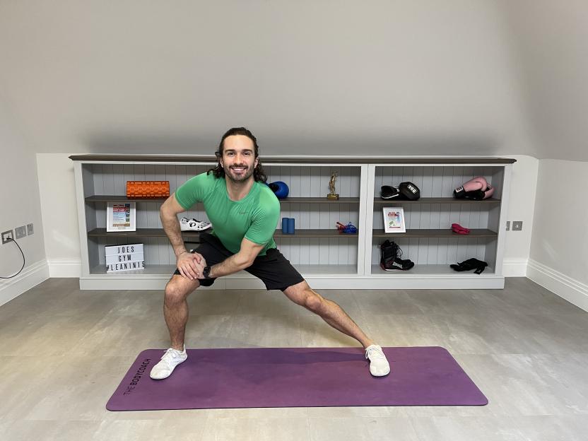 Joe Wicks doing a side lunge on a purple yoga mat in his home gym.