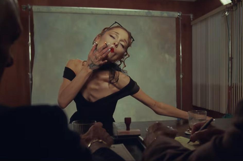 Ariana Grande with glasses on her head, dressed in black, holding a lit cigarette while leaning back in a chair.