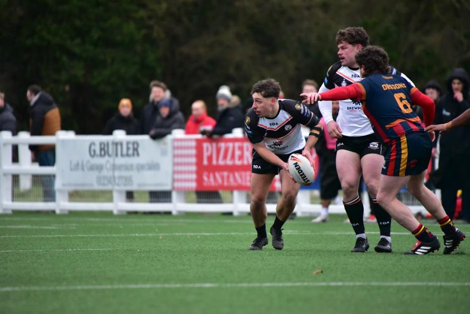 A rugby player in a black, white, and orange jersey holding a ball, preparing to pass while two other players stand nearby.