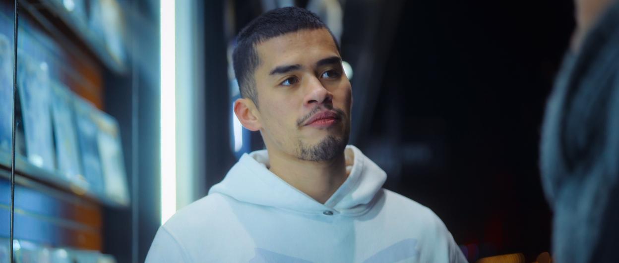 A young man with short dark hair and a light beard, wearing a white hoodie, looks to his right.