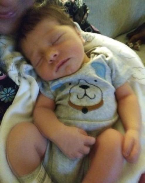 A baby boy in a bear onesie sleeping on a white blanket.