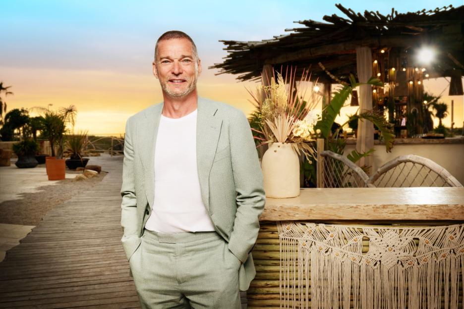 A smiling man in a light green suit standing on a boardwalk with a beach bar and sunset in the background.