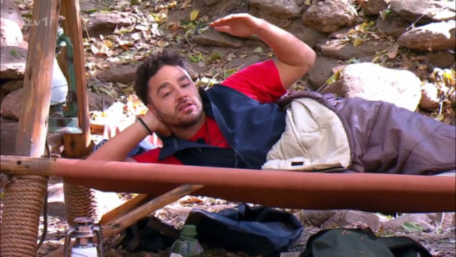 Adam Thomas lying on a cot in a red shirt and dark vest, looking upset.