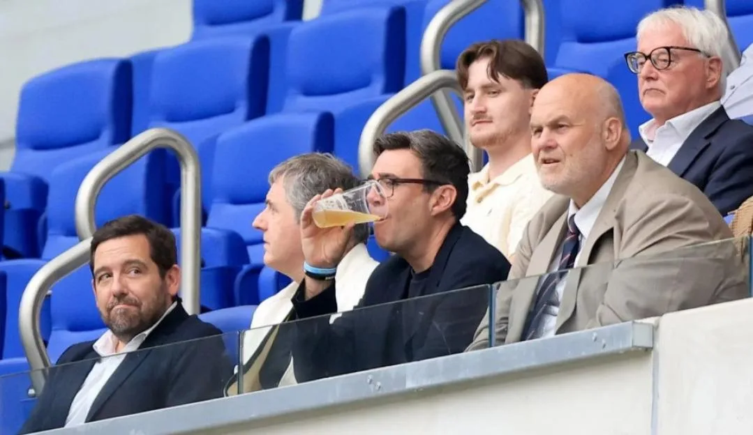 Andy Burnham watches from the stands, drinking a beverage.