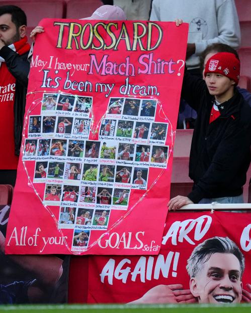 A football fan holds a red sign asking Leandro Trossard for his match shirt, with photos of Trossard in a heart shape.