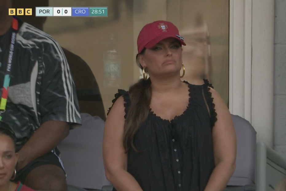 A woman in a red hat with the Portuguese emblem, sitting in the stands at a football match between Portugal and Croatia.