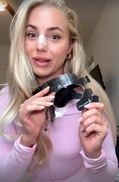 A woman with a bandage on her nose holds up a protective face shield.