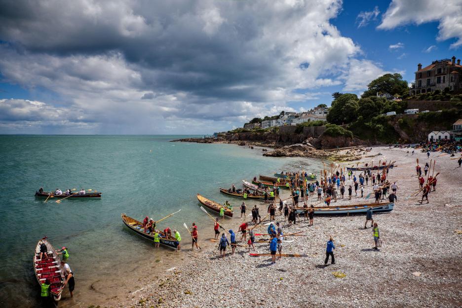 Boats and people on the rocky Breakwater Beach in Brixham, Devon, preparing for the Gig Regatta.
