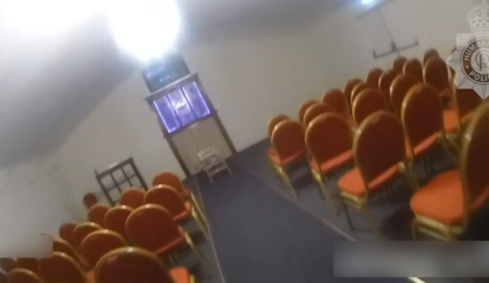 A room with rows of orange chairs facing a screen at the front, with a police badge watermark in the top right.