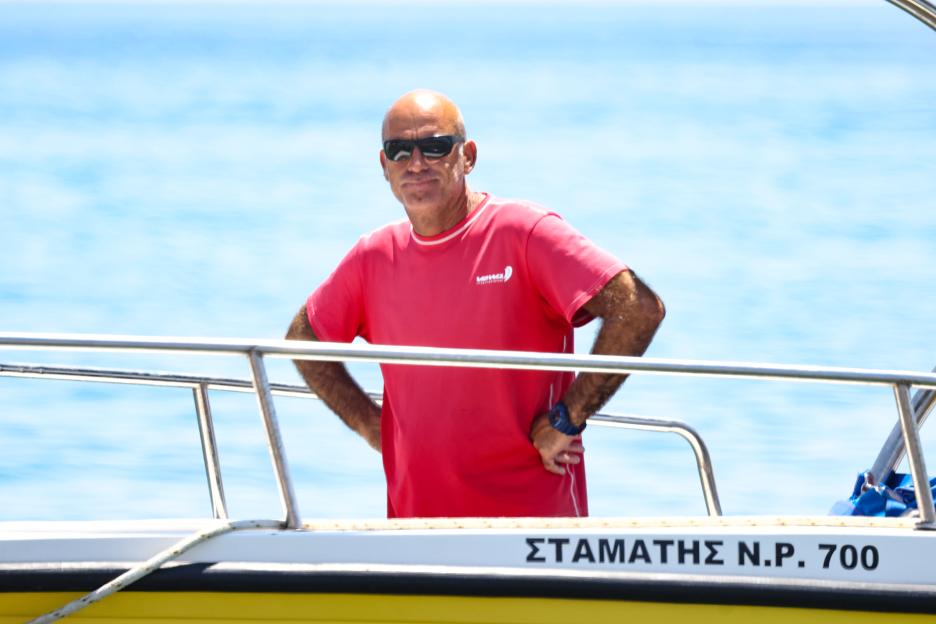 Nikos Mallios, wearing a red shirt and sunglasses, stands on a boat with the blue sky and sea behind him.
