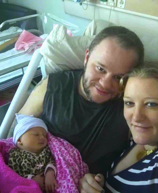 A man, woman, and sleeping infant posing for a picture in a hospital room.