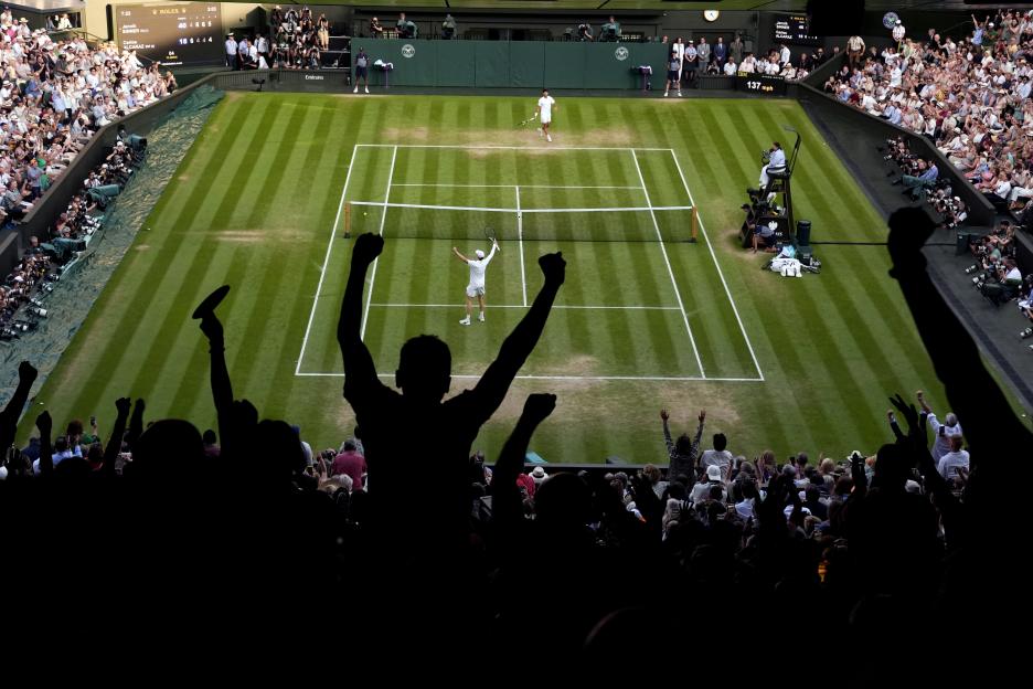 Jannik Sinner celebrates victory over Carlos Alcaraz on a tennis court, seen from a high angle with silhouetted spectators cheering in the foreground.