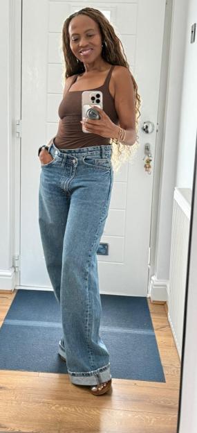 A woman with long braids wearing a brown tank top and blue jeans smiles while taking a mirror selfie.