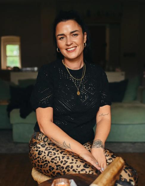 A smiling woman with dark hair and tattoos sitting in a living room, wearing a black top and leopard print pants.