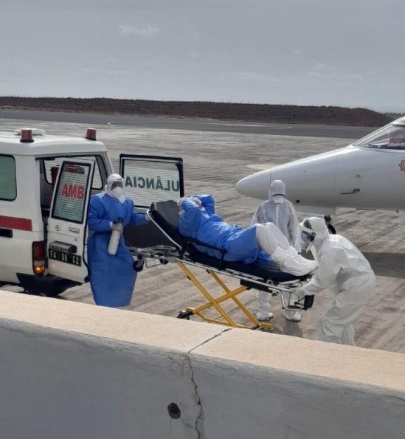 Medical personnel in hazmat suits transferring a patient on a stretcher from an ambulance to an air ambulance at an airport.