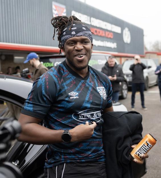 KSI wearing a Dagenham & Redbridge football jersey and holding a can of Prime Hydration drink.