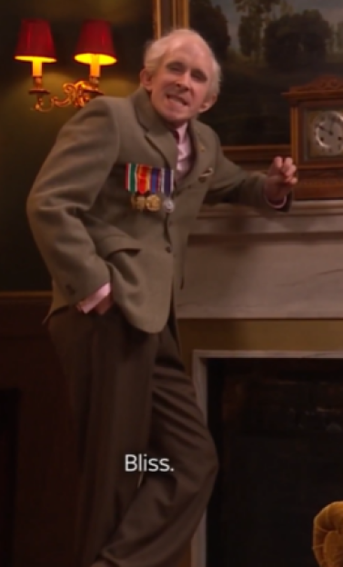 Elderly man with medals on his jacket leaning against a mantelpiece.