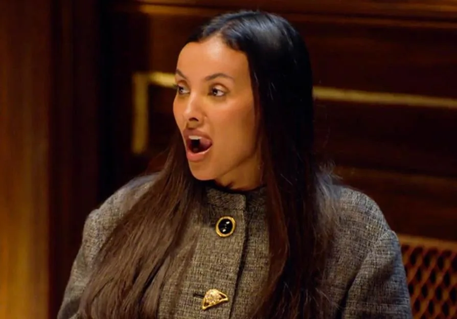 Maya Jama looking shocked in a dark tweed jacket with gold buttons.