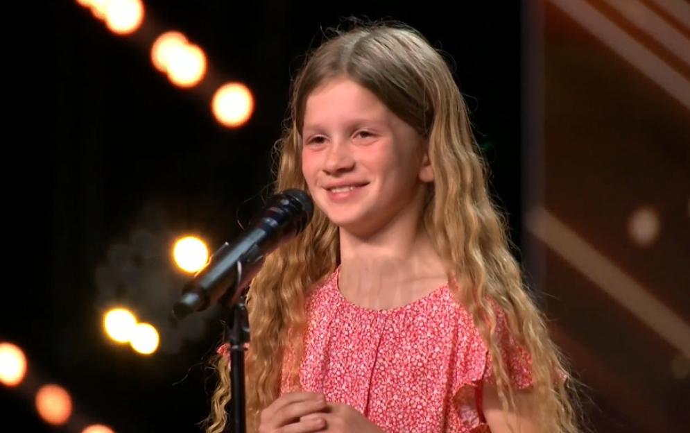 A young girl with long, wavy blonde hair smiles at a microphone, wearing a red and white floral top.