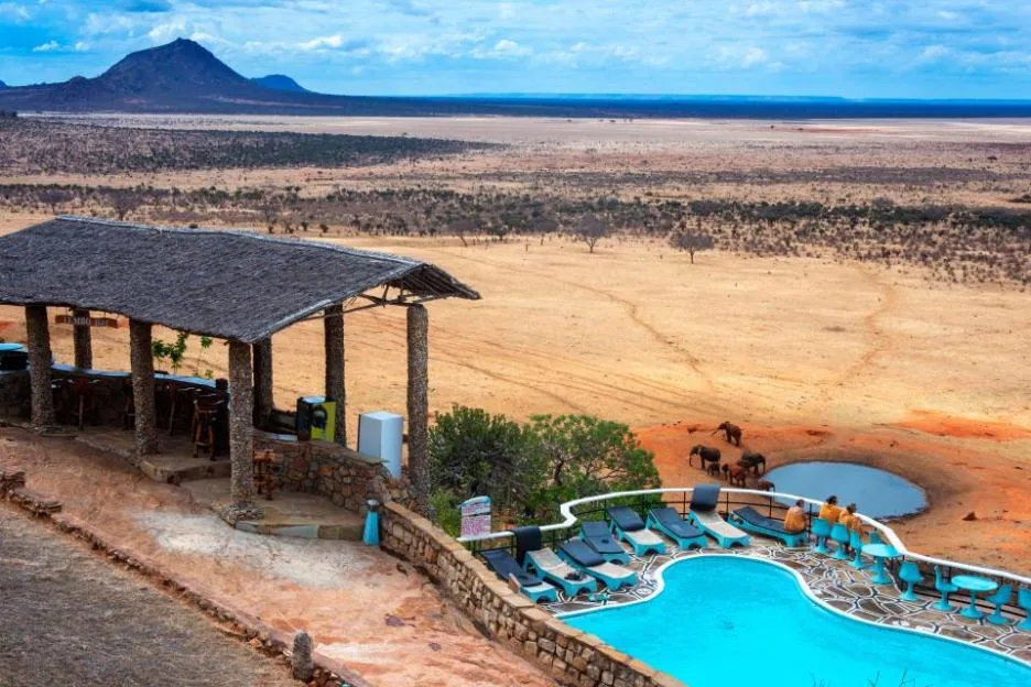 Guests at Voi Safari Lodge overlooking the savanna, a swimming pool, and elephants at a waterhole in Tsavo East National Park, Kenya.