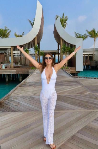 Elizabeth Hurley posing on a boardwalk over water, wearing a white monokini and white sheer lace pants.