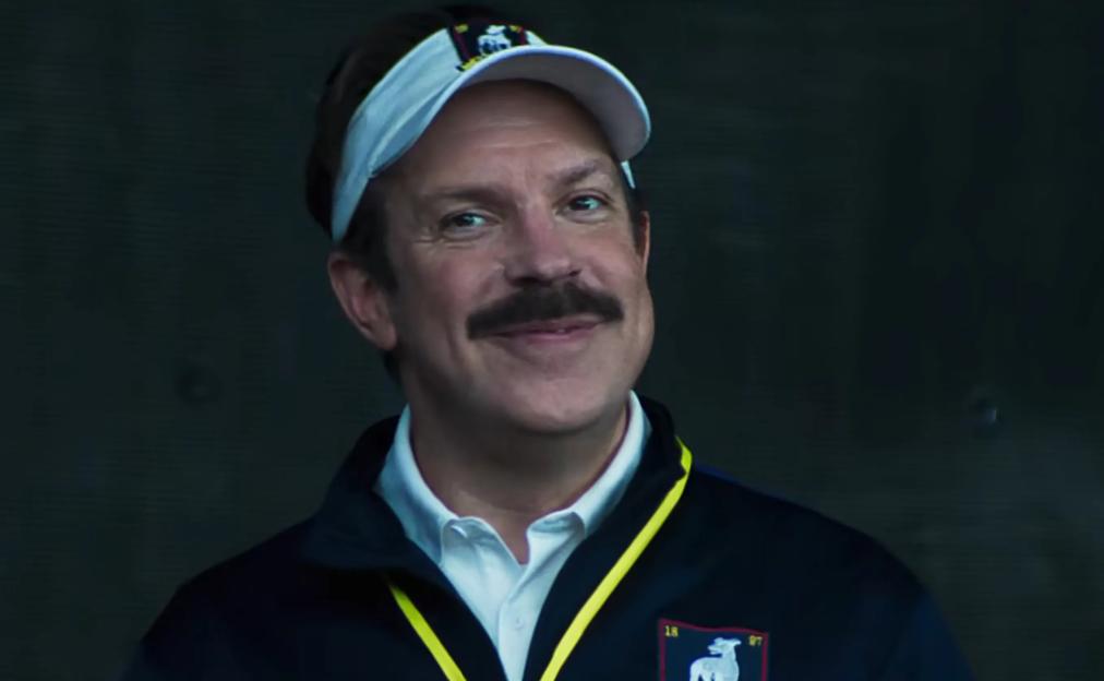 Actor Jason Sudeikis as Ted Lasso, wearing a white visor and a navy jacket with a yellow-collared polo shirt, smiling at the camera.