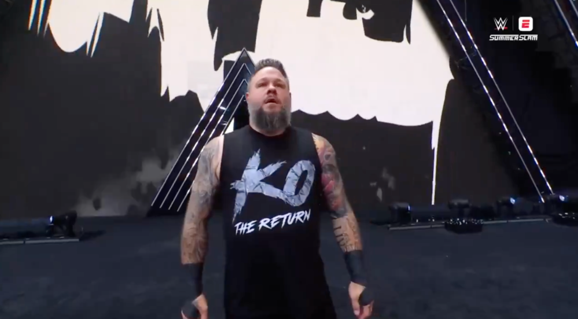 Kevin Owens wearing a black t-shirt that says "KO THE RETURN" on a stage.