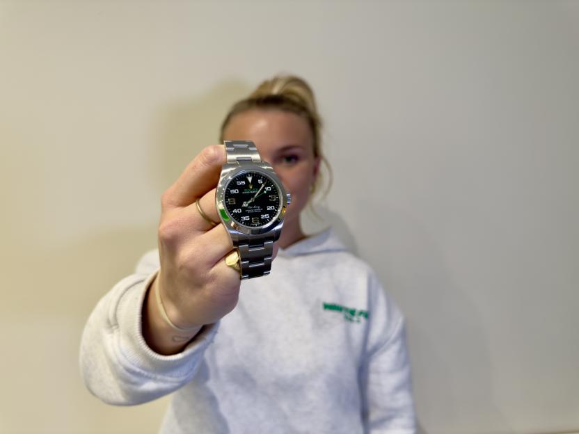 A person holding up a Rolex Air-King watch with a black face and stainless steel band, featuring green accents and the Rolex crown logo.