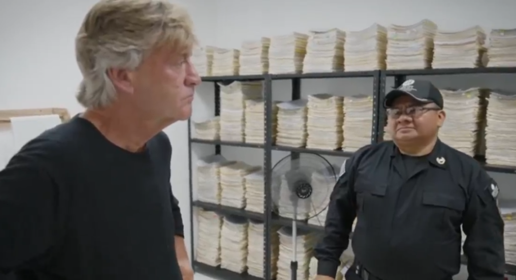 Two men in a storage room with shelves filled with stacks of paper.