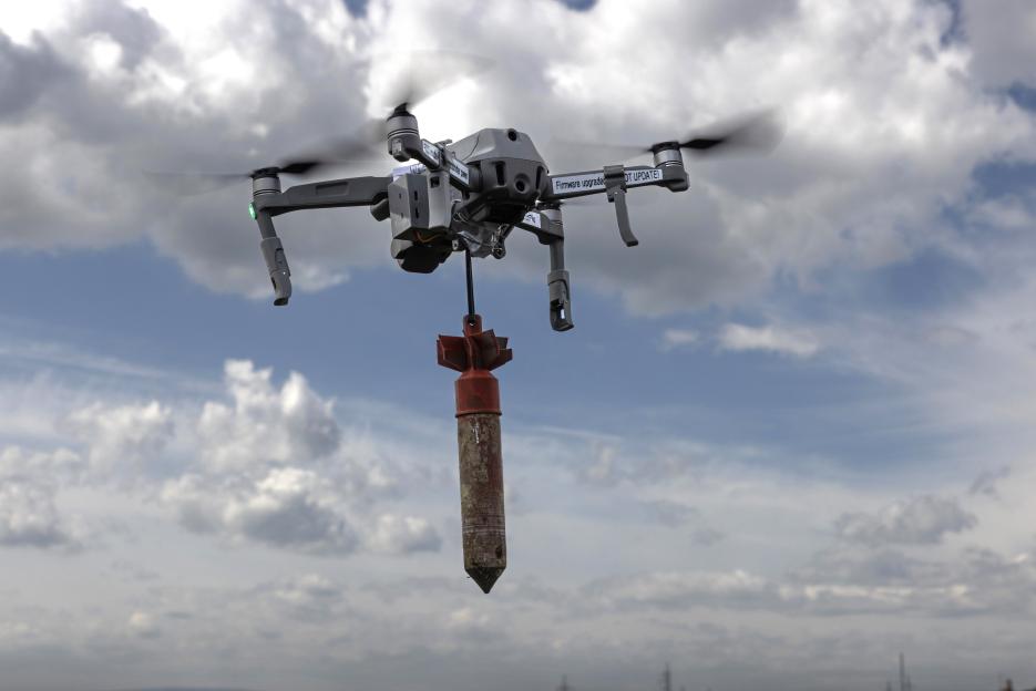 A drone with spinning propellers carries a bomb against a cloudy sky.