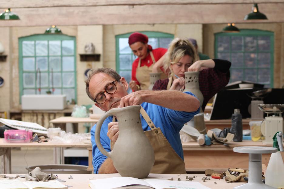 A man in glasses wearing an apron works on a clay pitcher, while two women work on smaller clay vases in the background.
