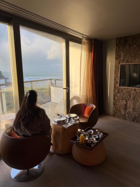A woman with long dark hair sits in a chair facing a window, looking out at the ocean.