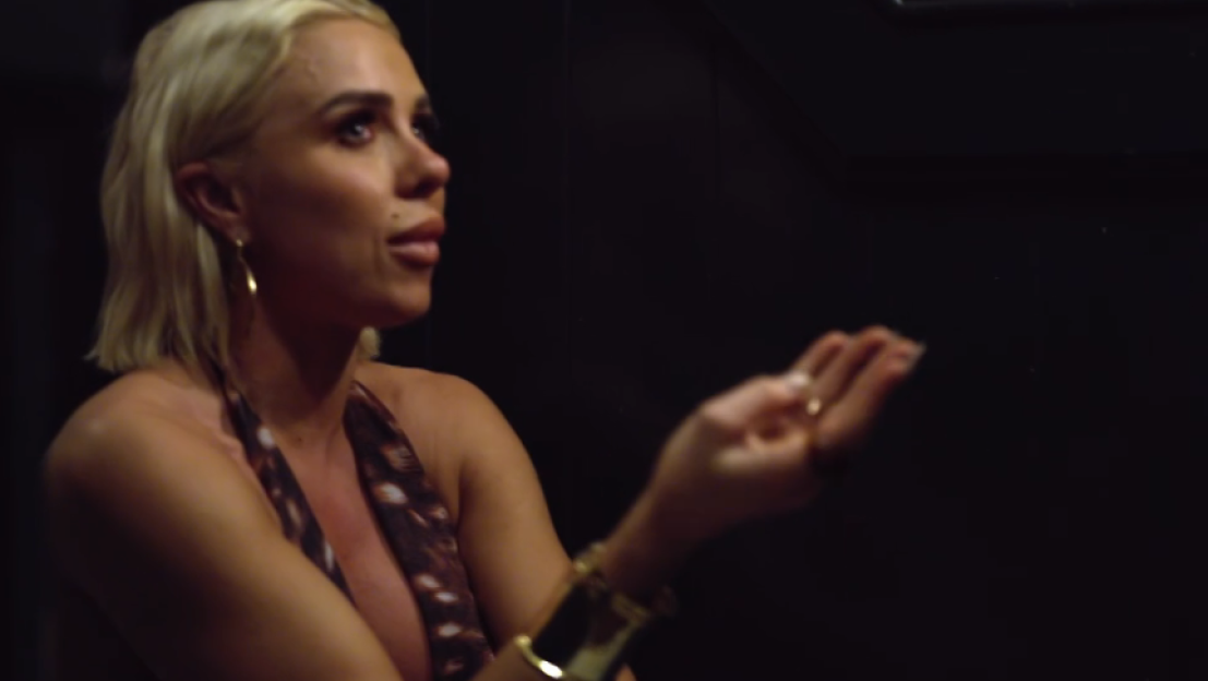 A woman with blonde hair and a patterned top, wearing a cuff bracelet, speaks with an open hand gesture.