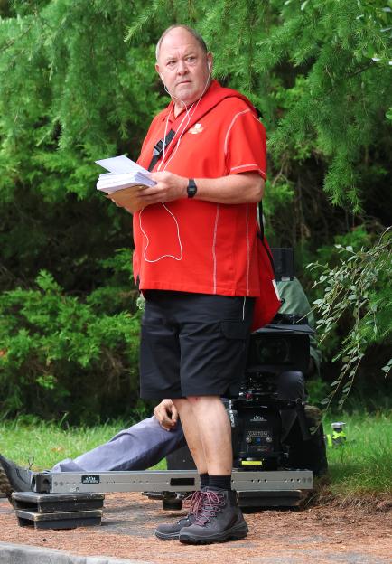 Mark Addy filming for "The Postman" in Bristol, wearing a red shirt, black shorts, and headphones, holding a stack of envelopes.