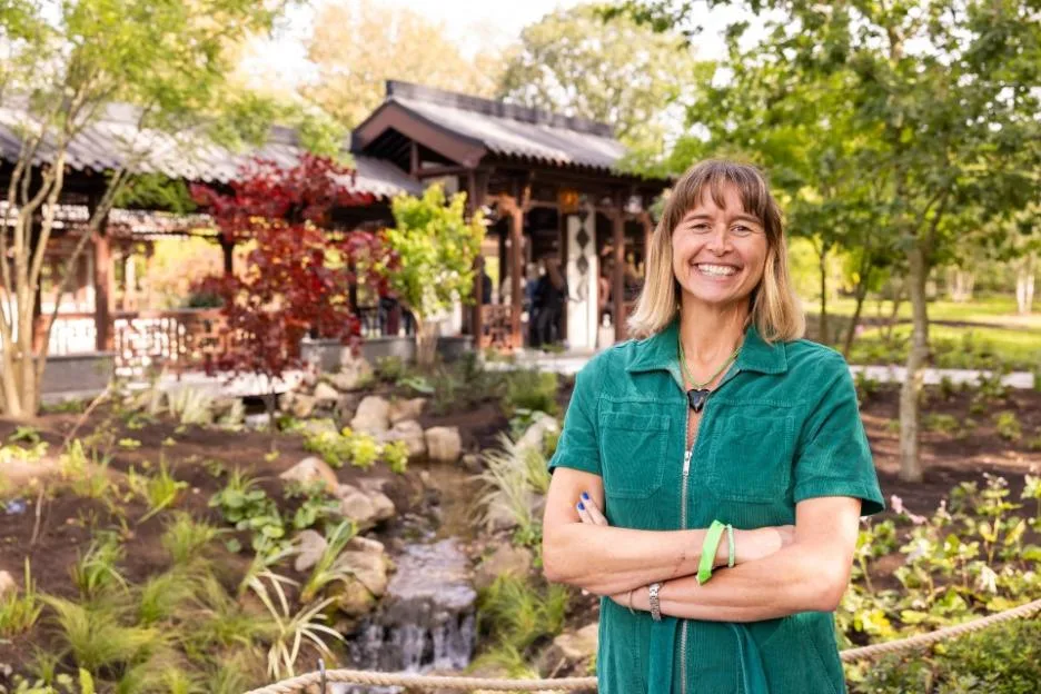 A smiling woman with shoulder-length hair stands with her arms crossed in front of an outdoor garden featuring a small stream and a traditional Asian pavilion.