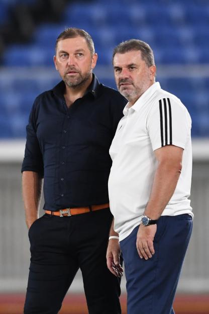 Two male soccer coaches, one in a dark blue shirt and one in a white polo, stand on the sidelines.