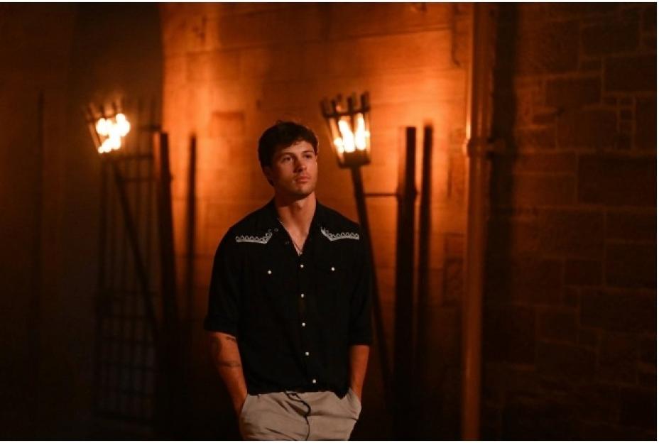 Rob Rausch from "The Traitors" looking upward, standing in an orange-lit medieval-style stone room with torch-like lights.