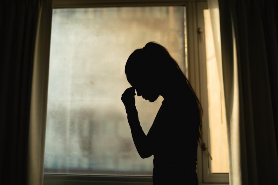 Silhouette of a distressed woman with her head in her hand, standing in front of a window.