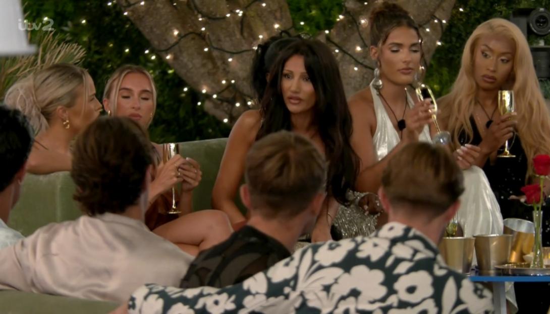 Female contestants with drinks seated on a couch, facing three male contestants.