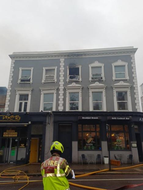 A firefighter stands before a multi-story building with fire damage and smoke stains around a broken window on an upper floor.