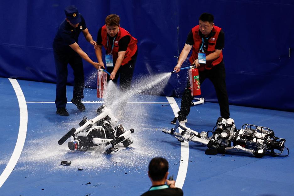 Two staff members use fire extinguishers on a humanoid robot that caught fire after a race, while another damaged robot lies nearby.