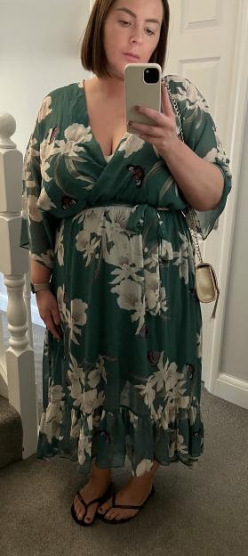 Amy Salmon posing in a green floral dress with a golden purse and flip-flops, holding a phone for a mirror selfie.