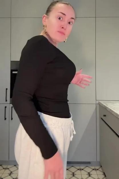 A woman in a black top and white sweatpants holds her stomach, showing excess skin.